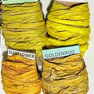 K&ouml;nnte beinhalten: Vier Spulen mit geknittertem Seidenband in Gelb- und Goldt&ouml;nen. Jede Spule ist mit Text beschriftet: Lemonade, Lemon, Sunflower und Goldenrod. Die B&auml;nder sind um Pappkerne gewickelt und weisen eine texturierte, ausgefranste Optik auf.