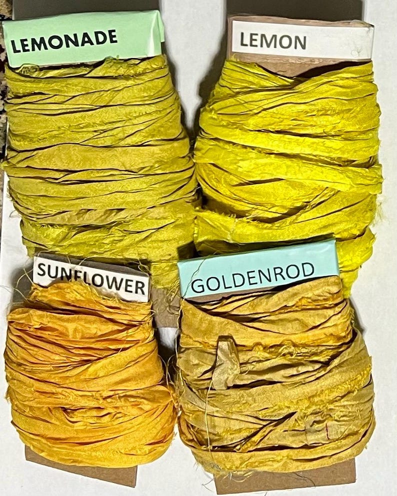 K&ouml;nnte beinhalten: Vier Spulen mit geknittertem Seidenband in Gelb- und Goldt&ouml;nen. Jede Spule ist mit Text beschriftet: Lemonade, Lemon, Sunflower und Goldenrod. Die B&auml;nder sind um Pappkerne gewickelt und weisen eine texturierte, ausgefranste Optik auf.