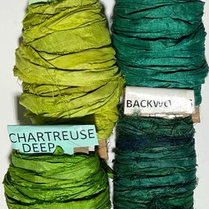 K&ouml;nnte beinhalten: Vier Seidenbandspulen in verschiedenen Gr&uuml;nt&ouml;nen. Die Etiketten tragen die Aufschriften Granny Smith, Ocean, Chartreuse Deep und Backwoods. Jede Spule besteht aus strukturiertem, zerknittertem Stoff, der um einen Pappkern gewickelt ist.