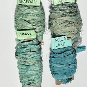 K&ouml;nnte beinhalten: Vier Spulen aus geknittertem Seidenband in Gr&uuml;n- und Blaut&ouml;nen. Die Etiketten tragen die Aufschriften SEAFOAM, AGAVE, ALOE und AQUA LAKE. Jede Spule besteht aus einem strukturierten Band mit ausgefransten Kanten, das um einen Pappkern gewickelt ist.