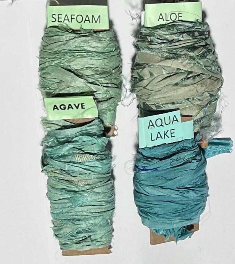 K&ouml;nnte beinhalten: Vier Spulen aus geknittertem Seidenband in Gr&uuml;n- und Blaut&ouml;nen. Die Etiketten tragen die Aufschriften SEAFOAM, AGAVE, ALOE und AQUA LAKE. Jede Spule besteht aus einem strukturierten Band mit ausgefransten Kanten, das um einen Pappkern gewickelt ist.