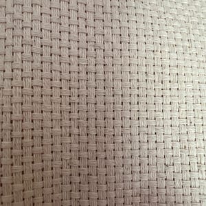 May include: A close-up view of off-white Aida cloth fabric featuring a distinct, uniform grid pattern created by the woven threads. The material displays a consistent, open-weave structure with visible square holes, commonly used as a base for needlepoint.
