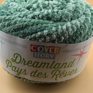 May include: A skein of soft, plush, variegated green and white yarn wrapped in a paper label. The label features the text COVER STORY, Dreamland, and Pays des Rêves, along with stylized starburst graphics in red, yellow, and white on a white background.