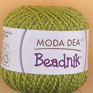 Pode incluir: Uma bola de fio verde com uma etiqueta que diz "MODA DEA Beadnik". O fio tem uma aparência brilhante e texturizada. A etiqueta também indica que o fio tem 50 gramas e 94 metros.