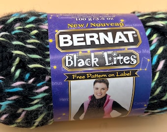 Könnte beinhalten: Ein Knäuel Bernat Black Lites Garn mit schwarzer Basis und Sprenkeln in Neonrosa, Blau und Gelb. Das violette Papieretikett zeigt den Text Bernat Black Lites, New/Nouveau, 100 g/3.5 oz sowie ein Foto einer Person, die einen Schal trägt.