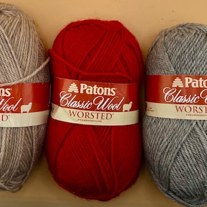Könnte beinhalten: Drei Knäuel Patons Classic Wool Worsted Garn in Hellbraun, leuchtendem Rot und meliertem Grau. Jedes Knäuel hat eine rot-weiße Papierbanderole mit dem Markennamen Patons, dem Text Classic Wool Worsted und einem kleinen Schaf-Symbol.