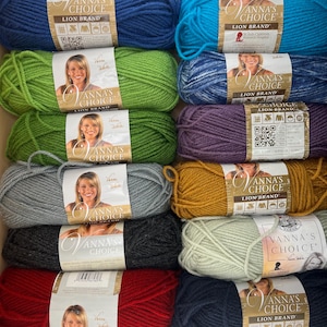 Puede incluir: Una colección de diez ovillos de lana Vanna's Choice Lion Brand en varios colores sólidos y jaspeados, incluyendo azul, verde, gris, negro, rojo, morado, dorado y verde claro, cada uno con una etiqueta de papel con el retrato de Vanna White.