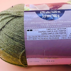 May include: A ball of olive green yarn with a label. The label indicates the yarn is 85% acrylic and 15% nylon, with 678 yards (620 meters) and 3.5 oz (100 g). The label also mentions instructions for a simple hat with a pom.