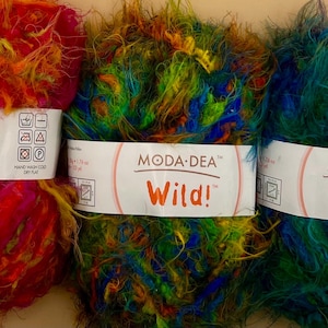 Pode incluir: Três novelos de lã coloridos com a etiqueta "MODA-DEA Wild!". As cores incluem vermelho, laranja, verde e azul. Perfeitos para tricô ou crochê. A lã é macia e fofa.