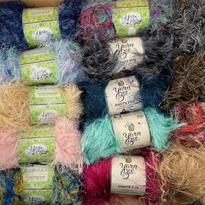 Puede incluir: Una colección de ovillos de lana Yarn Bee Haute Fur en varios colores, incluyendo azul, rosa, verde azulado, magenta, bronceado y negro. Cada ovillo tiene una etiqueta de papel con la marca, la línea de producto e instrucciones para agujas de 9 mm.