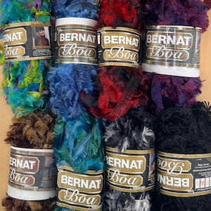 May include: A collection of Bernat Boa yarn in various colors, including teal, blue, red, purple, brown, and black. Each skein is labeled with the brand name and product details. The yarn has a fluffy, feather-like texture, ideal for crafting.