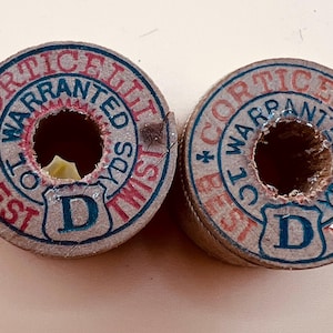 May include: Two vintage spools of thread with circular labels. The labels are tan with red and blue text that reads "Corticelli Warranted Best Twist". Each spool has a large center hole and the letter "D" in a shield.