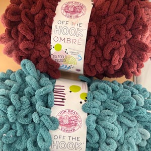 May include: Two skeins of Lion Brand Off The Hook Ombré yarn, one in deep burgundy and one in teal. Each skein features a white paper label with the text Lion Brand Yarns, Off The Hook Ombré, and No Tools Needed, decorated with green and purple dots.