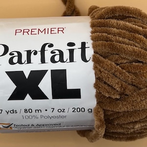 Puede incluir: Ovillo de lana marrón Premier Parfait XL. La etiqueta dice "87 yds / 80 m - 7 oz / 200 g - 100% Poliéster". La lana es un material suave y afelpado, ideal para diversos proyectos de manualidades.