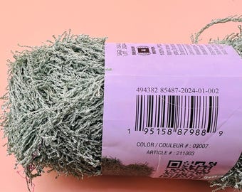 Può includere: Un gomitolo di filato testurizzato verde salvia con un'etichetta di carta viola chiaro avvolta attorno al centro. L'etichetta mostra un codice a barre, il numero 494382 85487-2024-01-002, il colore 03007, l'articolo 211003 e un codice QR.