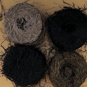 May include: Four balls of yarn in shades of black, gray, and gold. The yarn has a textured, fringed appearance, with strands extending outwards. The balls are arranged against a light brown background, showcasing the yarn's colors and textures.