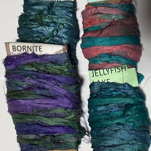 K&ouml;nnte beinhalten: Vier Spulen mit strukturiertem Seidenband in verschiedenen Farben. Die obere linke Spule ist mit DENIM in Dunkelblau beschriftet, die untere linke mit BORNITE in Lila und Gr&uuml;n, die obere rechte mit CORAL BELL in Pink und T&uuml;rkis und die untere rechte mit JELLYFISH LAKE in T&uuml;rkis.