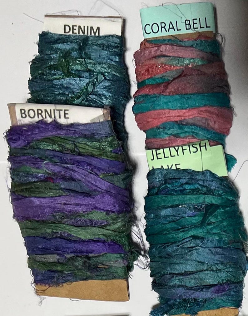 K&ouml;nnte beinhalten: Vier Spulen mit strukturiertem Seidenband in verschiedenen Farben. Die obere linke Spule ist mit DENIM in Dunkelblau beschriftet, die untere linke mit BORNITE in Lila und Gr&uuml;n, die obere rechte mit CORAL BELL in Pink und T&uuml;rkis und die untere rechte mit JELLYFISH LAKE in T&uuml;rkis.