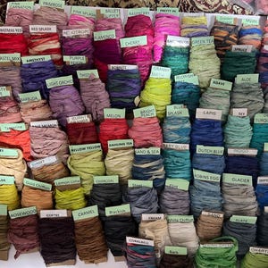 May include: A collection of numerous spools of hand-dyed silk ribbon, each wrapped around a cardboard tag labeled with a specific color name such as Pink Carnation, Raspberry, Green Tea, and Charcoal, arranged in a grid of various hues.