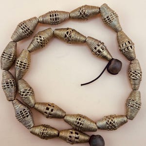 May include: A strand of gold-coloured metal beads. The beads are cylindrical and have a woven pattern. The beads are strung on a black cord with a dark brown bead at each end.