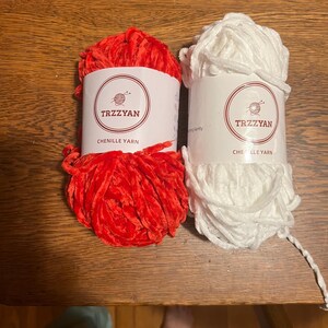 Könnte beinhalten: Zwei Knäuel Chenille-Garn, eines rot und eines weiß, liegen auf einer Holzoberfläche. Jedes Knäuel hat ein Etikett mit dem Text "TRZZYAN" und "CHENILLE YARN". Das Garn wirkt weich und flauschig, ideal für verschiedene Bastelprojekte.