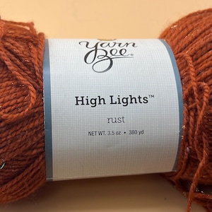Può includere: Un gomitolo di filato Yarn Bee High Lights color ruggine. L'etichetta recita "High Lights" e "rust". Il filato è da 99 g e 347 m. Il filato ha una leggera lucentezza.