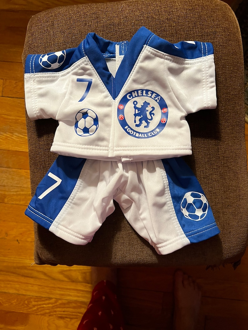 Puede incluir: Camiseta de f&uacute;tbol blanca y azul con el n&uacute;mero 7 y el logotipo del Chelsea Football Club. La camiseta se combina con pantalones cortos blancos y azules a juego.