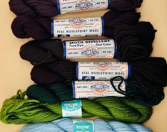 May include: Seven skeins of Hiawatha and Bucilla needlepoint and crewel wool yarn arranged in a stack. The colors include dark purple, black, dark green, olive green, and light blue. Each skein features a paper label with brand text.