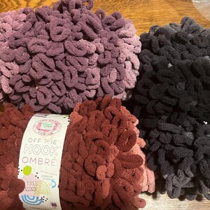 May include: Three bundles of Off The Hook Ombre yarn in shades of purple, black, and red. The yarn is thick and looped, with a label visible on one bundle. The label reads "Off The Hook Ombre".