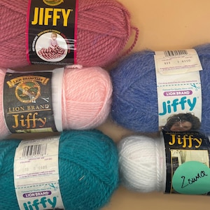 May include: Assortment of Jiffy brand yarn skeins in various colors, including pink, blue, teal, and white. Each skein has a label with the Lion Brand logo and product information. The yarn is soft and fluffy, ideal for knitting and crochet projects.