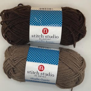 Könnte beinhalten: Zwei Knäuel Stitch Studio by Nicole Studio Basic Garn. Eines ist dunkelbraun, das andere hellbraun. Die Etiketten sind weiß mit blauen Punkten und dem Markennamen.