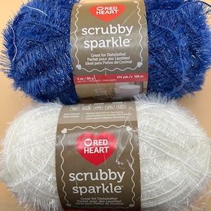May include: Two skeins of Red Heart Scrubby Sparkle yarn, one in royal blue and one in white. Each skein features a textured, metallic-flecked polyester fibre and a central paper label displaying the brand name, product name, and weight of 85 g.