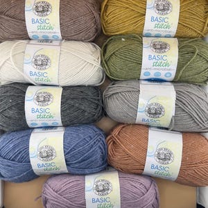 Puede incluir: Una colección de madejas de hilo de colores en una caja de madera. El hilo es de varios colores, incluyendo marrón, amarillo, verde, gris, azul y rosa. Cada madeja tiene una etiqueta que dice "BASIC stitch anti-microbial".