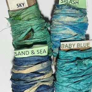 K&ouml;nnte beinhalten: Vier Spulen aus zerknittertem Seidenband in Gr&uuml;n- und Blaut&ouml;nen. Jede Spule hat ein Papieretikett mit dem Text: SKY, BLUE SPLASH, SAND & SEA und BABY BLUE. Die B&auml;nder haben ein strukturiertes, ausgefranstes Aussehen und sind um Pappkerne gewickelt.