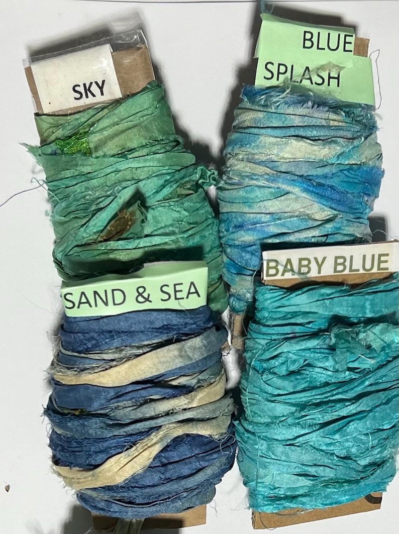 K&ouml;nnte beinhalten: Vier Spulen aus zerknittertem Seidenband in Gr&uuml;n- und Blaut&ouml;nen. Jede Spule hat ein Papieretikett mit dem Text: SKY, BLUE SPLASH, SAND & SEA und BABY BLUE. Die B&auml;nder haben ein strukturiertes, ausgefranstes Aussehen und sind um Pappkerne gewickelt.