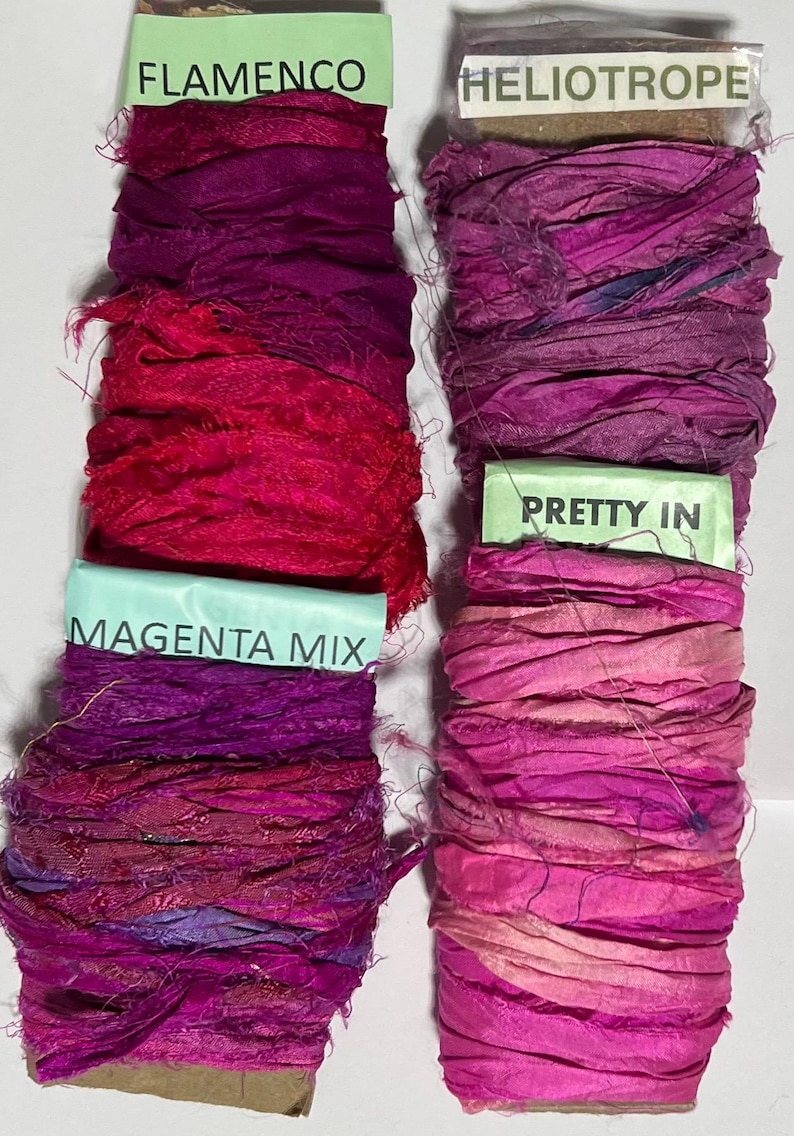 K&ouml;nnte beinhalten: Vier Seidenbandspulen in verschiedenen Rosa- und Violettt&ouml;nen. Die Etiketten tragen die Aufschriften Flamenco, Heliotrope, Magenta Mix und Pretty In. Jede Spule besteht aus ausgefranstem, strukturiertem Stoff auf einem Pappkern.