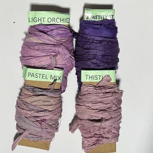 K&ouml;nnte beinhalten: Vier Spulen aus zerknittertem Seidenband in Violettt&ouml;nen, beschriftet mit Light Orchid, Amethyst, Pastel Mix und Thistle. Jede Spule ist mit strukturierten, handgef&auml;rbten Stoffstreifen mit ausgefransten Kanten auf Pappkarten aufgewickelt.