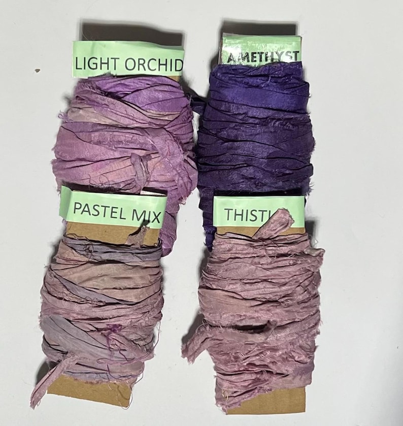 K&ouml;nnte beinhalten: Vier Spulen aus zerknittertem Seidenband in Violettt&ouml;nen, beschriftet mit Light Orchid, Amethyst, Pastel Mix und Thistle. Jede Spule ist mit strukturierten, handgef&auml;rbten Stoffstreifen mit ausgefransten Kanten auf Pappkarten aufgewickelt.