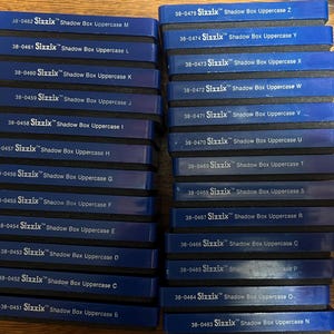 May include: A collection of blue Sizzix Shadow Box Uppercase letter dies, stacked in two columns. Each die is labeled with a letter from the alphabet, from B to Z, and a corresponding product number. The dies are for crafting and scrapbooking.