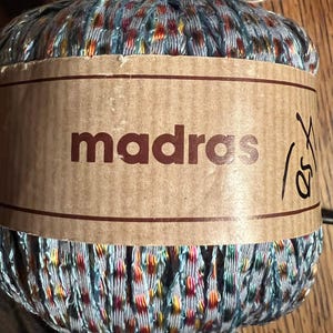 Puede incluir: Ovillo de hilo Madras con una etiqueta marrón. El hilo es una mezcla de azul, rojo, amarillo y naranja. La palabra "madras" está impresa en marrón en la etiqueta. Se ve un número escrito a mano.