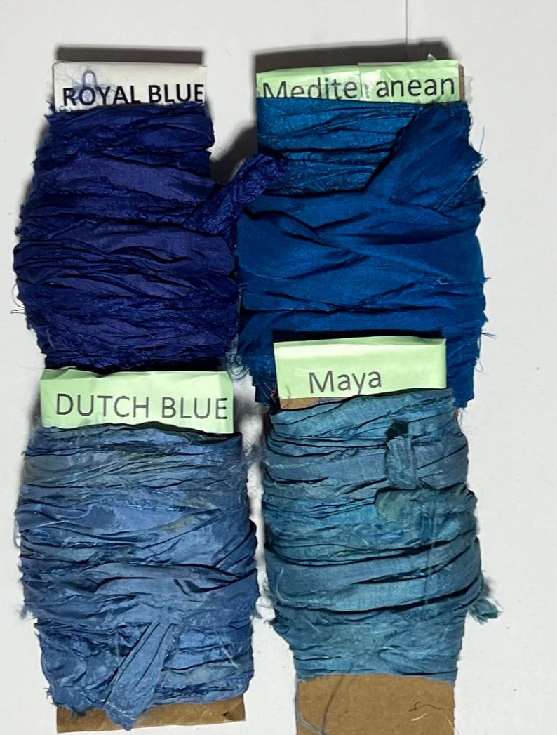 K&ouml;nnte beinhalten: Vier Spulen aus zerknittertem Seidenband in Blaut&ouml;nen. Die obere linke Spule ist mit ROYAL BLUE beschriftet, die obere rechte mit Mediterranean, die untere linke mit DUTCH BLUE und die untere rechte mit Maya auf hellgr&uuml;nem Papier.
