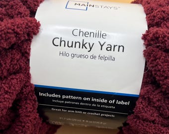 Può includere: Un gomitolo di filato ciniglia spesso color bordeaux Mainstays con un'etichetta di carta bianca. Il testo sull'etichetta recita Mainstays, Chenille Chunky Yarn, Includes pattern on inside of label, 31,7 yds (29 m) e 8 oz (226,8 g).
