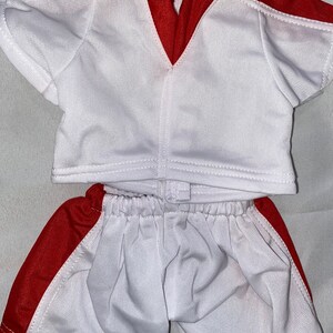 Puede incluir: Uniforme de f&uacute;tbol blanco y rojo para una mu&ntilde;eca. La camiseta tiene un cuello en V rojo y ribetes rojos en las mangas. Los pantalones cortos tienen una raya roja a los lados.