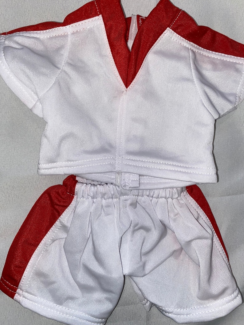 Puede incluir: Uniforme de f&uacute;tbol blanco y rojo para una mu&ntilde;eca. La camiseta tiene un cuello en V rojo y ribetes rojos en las mangas. Los pantalones cortos tienen una raya roja a los lados.