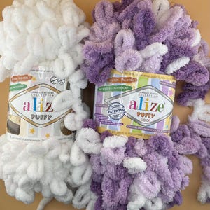 Puede incluir: Dos ovillos de hilo Alize Puffy, uno blanco y otro morado. El embalaje indica "¡Sin agujas, sin ganchos!" y "Puffy Color". Ideal para tejer fácilmente.