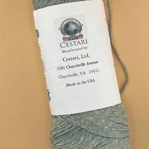 May include: A skein of light grey yarn with a white paper label. The label features a circular logo with a cross and the text Cestari, Manufactured by: Cestari, Ltd., 3581 Churchville Avenue, Churchville, VA 24421, Made in the USA, and care symbols.