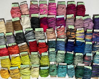 May include: A collection of numerous spools of crinkled silk ribbon in a wide variety of colors, each featuring a small paper label with a specific color name printed on it, arranged in a grid pattern to display the full spectrum of available shades.