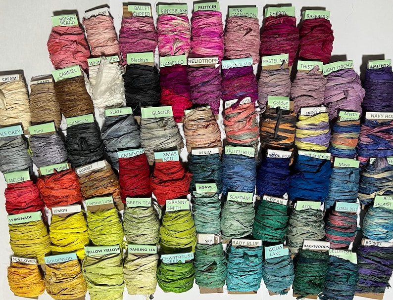 K&ouml;nnte beinhalten: Eine Sammlung zahlreicher Rollen aus geknittertem Seidenband in einer Vielzahl von Farben, jede mit einem kleinen Papieretikett versehen, auf dem der Farbname gedruckt ist, angeordnet in einem Raster, um das gesamte Farbspektrum zu zeigen.