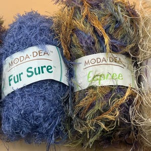 Pode incluir: Quatro novelos de fio Moda Dea em várias cores. Os nomes dos fios são Jai Alai, Fur Sure, Espree e Prima. O fio é fofo e texturizado, com uma mistura de azul, castanho e bege.