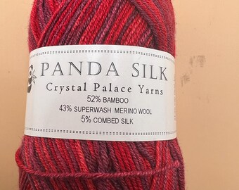 May include: A skein of Panda Silk yarn by Crystal Palace Yarns in variegated shades of red and purple. The white label displays the text: PANDA SILK, Crystal Palace Yarns, 52% BAMBOO, 43% SUPERWASH MERINO WOOL, 5% COMBED SILK.